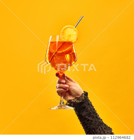 Female hand hold delicious Aperol Spritz cocktail garnished with slice of orange against orange background. 112964682