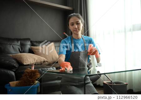 Millennial woman wearing gloves wiping dust and cleaning glass table in living room Millennial woman wearing gloves wiping dust and cleaning glass table in living room 112966520