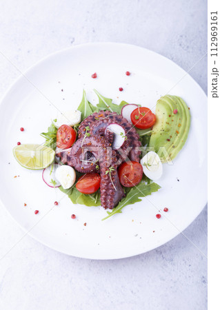 Warm salad with octopus, cherry tomatoes, avocado, arugula, quail eggs, radish and lime on a white plate. Traditional Mediterranean dish. Close-up, white background. 112969161