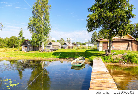 Old fishing plastic boat moored at a pier on the lake and traditional russian wooden baths on the shore in the summer 112970303