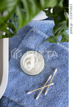 Opened cream jar on blue towels near basin and green monstera in bath, beauty routine 112972621