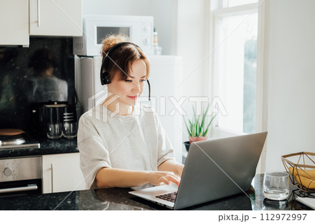 Remote work at home. Woman in headset working on Laptop at Kitchen Table in tiny flat. Student has online classes. Manager has video meeting. Customer support operator in telemarketing service 112972997
