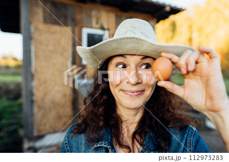 Portrait of funny joyful farmer woman holding fresh egg near her face. While enjoying farming, a female farmer holds a fresh egg Happy moment of collecting eggs 112973283