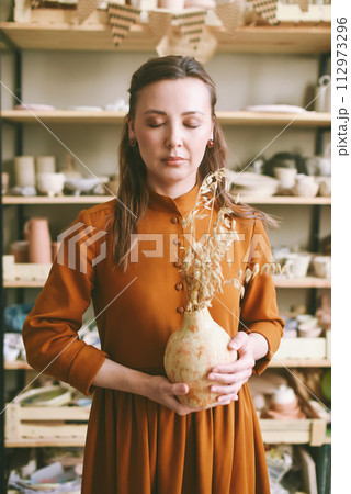 Creative Woman Potter with Clay Vase in Artisan Studio with eyes closed. Artistic woman enjoying her hobby, holding a unique piece of pottery in a well-lit studio 112973296
