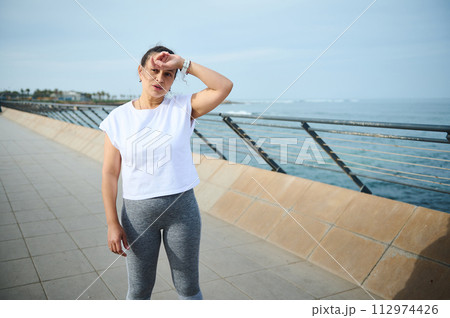 Exhausted young athletic woman runner wiping her forehead, standing on the bridge, relaxing after morning jog 112974426