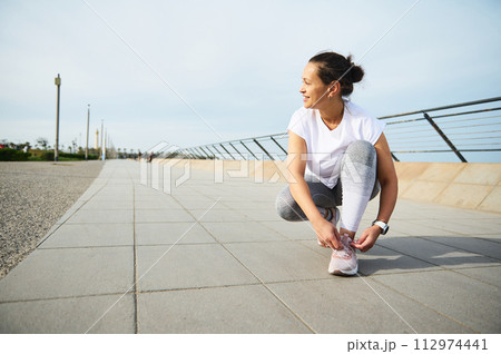 Young woman jogger tying laces on her pink sneakers, smiling looking away, getting ready for morning jog in the city Young woman jogger tying laces on her pink sneakers, smiling looking away, getting ready for morning jog in the city 112974441