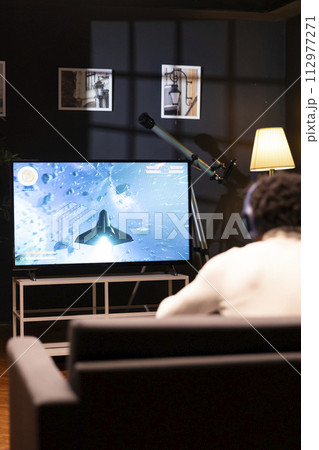 Gamer in front of widescreen smart TV watching SF videogame gameplay. African american man wearing headphones in home theatre to enjoy gaming content online displayed on television set 112977271