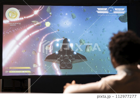 Man plays science fiction singleplayer game with overlay HUD showing health bars and minimap on ultrawide smart TV screen. Player enjoying high quality graphics in cloud gaming streamed videogame 112977277