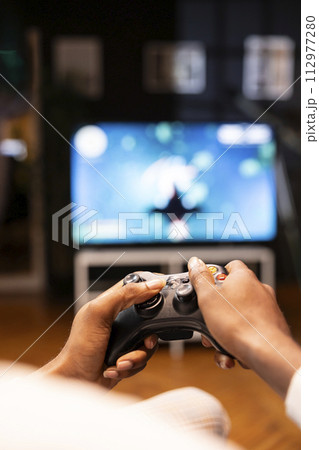 Focus shot on man holding controller in apartment, playing videogames on smart TV display in blurry background. Gamer on sofa using joypad to participate in game on console attached to television set 112977280
