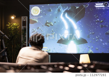 BIPOC man plays science fiction singleplayer game with overlay HUD showing health bars and minimap on huge TV screen. Player enjoying high quality ray tracing graphics in console videogame 112977281