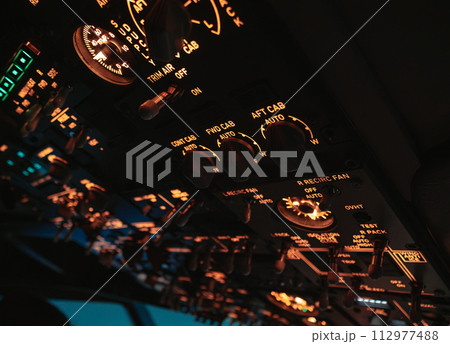 A close-up view of an aircrafts cockpit dashboard highlights an array of lit dials and switches, glowing in the subdued light 112977488