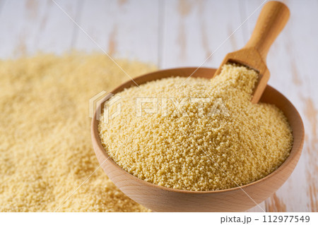 Organic couscous are scattered out of the wooden bowl on a light table, selective focus. Organic couscous are scattered out of the wooden bowl on a light table, selective focus. 112977549