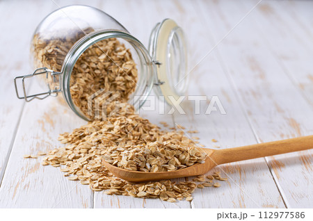 Raw oatmeal flakes in wooden scoop and in glass storage jar on a white wooden table. 112977586
