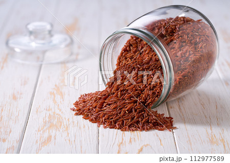 Raw red rice spill out of a glass storage jar on a white wooden table. 112977589