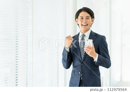 スマホを持ってガッツポーズするスーツ姿の若いアジア人男性ビジネスマン 112979564
