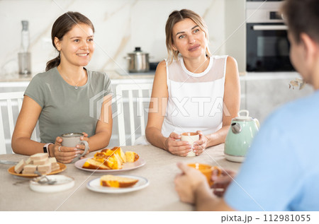 Smiling woman and girl receiving male guest over cup of tea at home 112981055