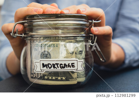 Unrecognizable woman holding Saving Money In Glass Jar filled with Dollars banknotes. MORTGAGE transcription in front of jar. Managing personal finances extra income for future insecurity 112981299