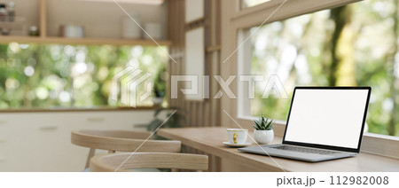 A laptop computer mockup on a wooden dining table against the window in a neutral modern kitchen. 112982008