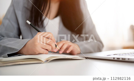 A cropped shot of a businesswoman focused on writing or taking notes in the notebook with a pencil. 112982116