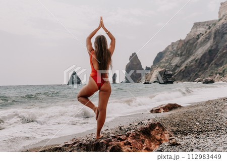 Woman sea yoga. Back view of free calm happy satisfied woman with long hair standing on top rock with yoga position against of sky by the sea. Healthy lifestyle outdoors in nature, fitness concept. Woman sea yoga. Back view of free calm happy satisfied woman with long hair standing on top rock with yoga position against of sky by the sea. Healthy lifestyle outdoors in nature, fitness concept. 112983449