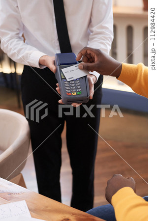 Vertical shot of unrecognizable Black man paying for order in restaurant with credit card tapping it onto terminal waiter holding Vertical shot of unrecognizable Black man paying for order in restaurant with credit card tapping it onto terminal waiter holding 112984420