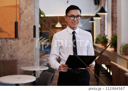 Medium portrait of young Asian restaurant manager wearing eyeglasses holding menu smiling at camera, copy space Medium portrait of young Asian restaurant manager wearing eyeglasses holding menu smiling at camera, copy space 112984567