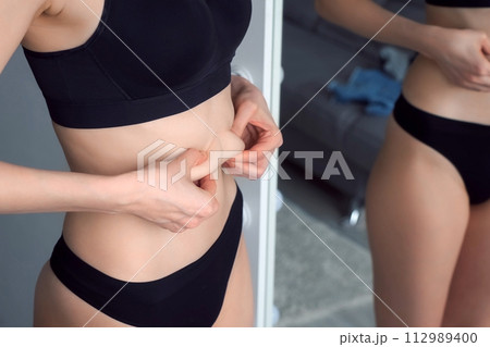 A young thin woman in black underwear critically examines her figure in mirror. 112989400