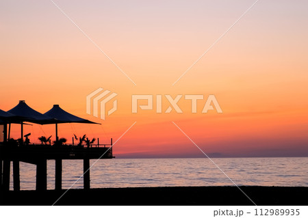 Tropical ocean beach with cafe on beach at dark sunset with purple orange sky. Tropical ocean beach with cafe on beach at dark sunset with purple orange sky. 112989935