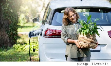 Woman with plant in pot next to a charging electric car in the yard of a country house Woman with plant in pot next to a charging electric car in the yard of a country house 112990417