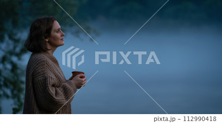 A woman in a sweater on the bank of a foggy river in the evening A woman in a sweater on the bank of a foggy river in the evening 112990418