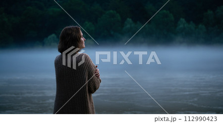 A woman in a sweater on the bank of a foggy river in the evening A woman in a sweater on the bank of a foggy river in the evening 112990423