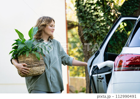 Woman with plant in pot next to a charging electric car in the yard of a country house 112990491