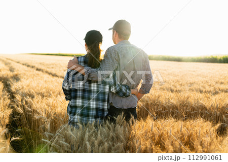 A couple of farmers in plaid shirts and caps stand embracing on agricultural field of wheat at sunset. 112991061