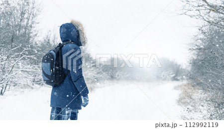 Back view of traveler in a blue jacket with a fur hood and a backpack on the background of a winter landscape during snowfall. 112992119