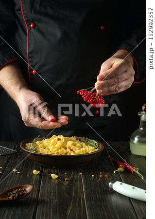 Adding viburnum berries in a cook hand to a plate of pasta. The chef at the kitchen table in the tavern is preparing a delicious lunch 112992357