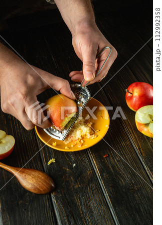 Close-up of a chef hands grating apples with a grater. Preparing applesauce on the kitchen table. Advertising space 112992358