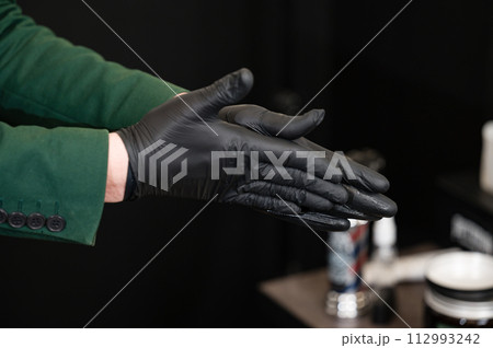 A barber in black gloves rubs wax and pomade for styling a clients hair. 112993242