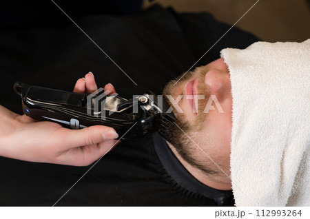 The clients head is covered with a towel during a beard trim. Shortening the length of the beard from the sides by the master for the client. The clients head is covered with a towel during a beard trim. Shortening the length of the beard from the sides by the master for the client. 112993264