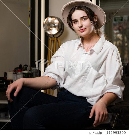 Portrait of a young hairdresser girl in a hat. A female barber is sitting in a chair and waiting for a client. Portrait of a young hairdresser girl in a hat. A female barber is sitting in a chair and waiting for a client. 112993290