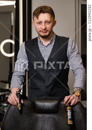 A barber in a shirt and vest stands near a barbers chair holding tools for haircuts. A barber in a shirt and vest stands near a barbers chair holding tools for haircuts. 112993292