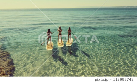 Three happy women stands on a sup boards. In the background, the ocean and the sunset 112995745