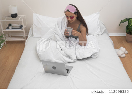 A woman, bed-bound with eye patches and coffee, navigates online work and remote studies seamlessly through her laptop from her home sanctuary. A woman, bed-bound with eye patches and coffee, navigates online work and remote studies seamlessly through her laptop from her home sanctuary. 112996326