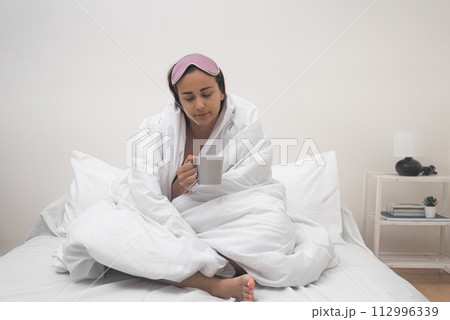 A weary woman, cocooned in a blanket, perches on her bed, clutching a coffee cup, a vivid depiction of insomnia's toll on her sleep.  112996339