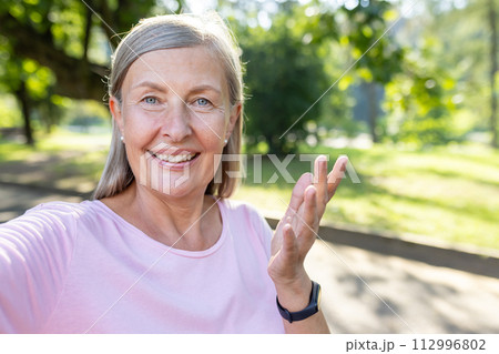 Close-up photo of an active senior gray-haired woman walking in the park, talking on the phone with a video camera, smiling and gesturing with her hands. 112996802