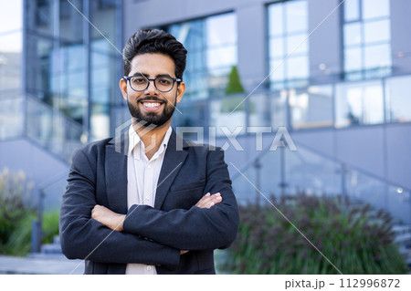 Portrait of a young successful and smiling Muslim male businessman standing outside an office building in a suit and glasses, crossing his arms and looking confidently at the camera. 112996872