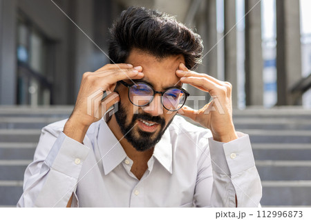 A close-up photo of a young Indian male office worker sitting outside a building on the steps and holding his head in his hands, feeling severe pain and pressure. A close-up photo of a young Indian male office worker sitting outside a building on the steps and holding his head in his hands, feeling severe pain and pressure. 112996873