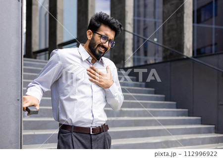 Contorted in pain, a young Indian male businessman stands by the wall of an office building and holds his hand over his heart. 112996922