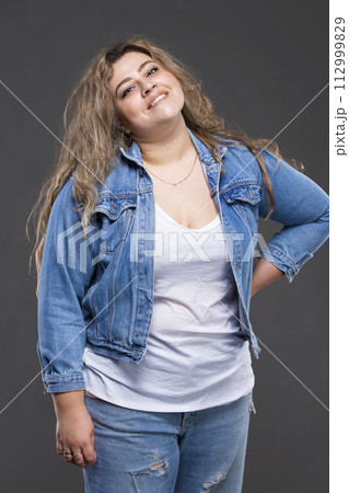 A beautiful fat woman in a denim jacket looks at the camera and smiles on a gray background. 112999829