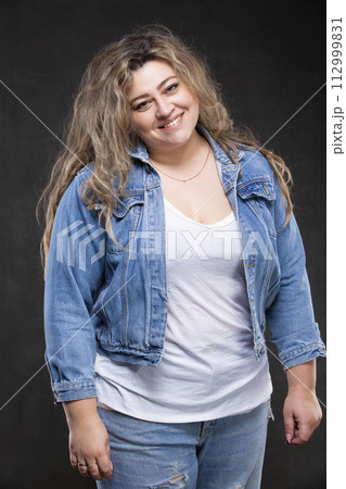 A beautiful fat woman in a denim jacket looks at the camera and smiles on a gray background. 112999831