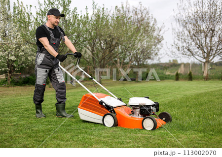 Professional gardener in protective apparel is mowing green grass lawn using modern gasoline cordless lawnmower at backyard. Seasonal landscaping design work. Blooming trees on background 113002070